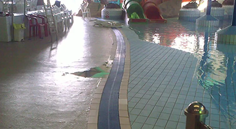 Indoor swimming pool area with a damaged floor section near the pool edge; water slides and pool chairs are visible in the background.