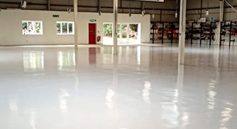A spacious, empty industrial warehouse interior with a highly polished white floor, white walls, large windows, shelves in the back, and several support columns. Bright lighting reflects off the clean, shiny surface.