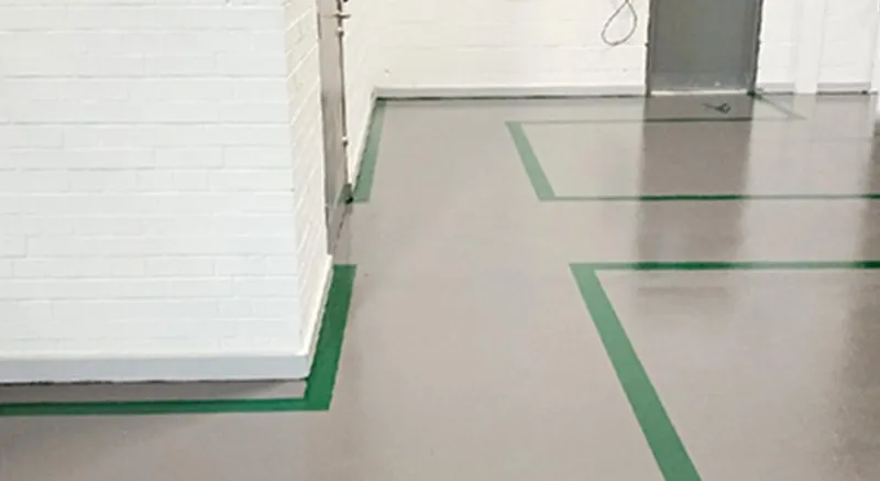 A gray floor with green lines marking pathways around white brick walls in an indoor setting, possibly a warehouse or industrial facility. A closed door and some wires are visible in the background.