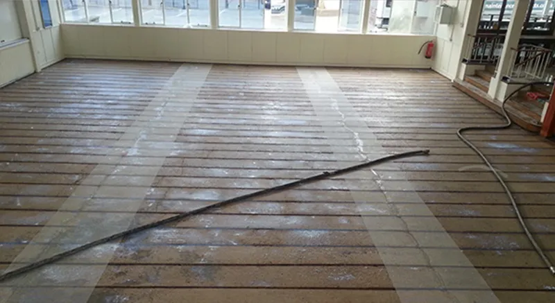 A large indoor room with exposed floorboards and several strips of light-colored material. A long black cable or hose lies diagonally across the floor. Sunlight enters through large windows in the background.