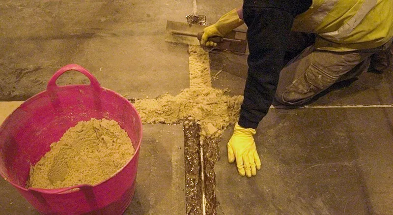 A person wearing yellow gloves uses a trowel to fill a crack in a concrete floor with sand or grout. A large pink bucket filled with sand sits nearby.