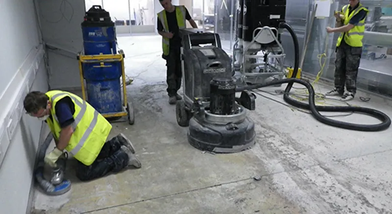 Three workers in yellow safety vests clean and polish a concrete floor using industrial equipment in an unfinished indoor space. One operates a floor polisher, another kneels cleaning the floor, and a third manages cables.