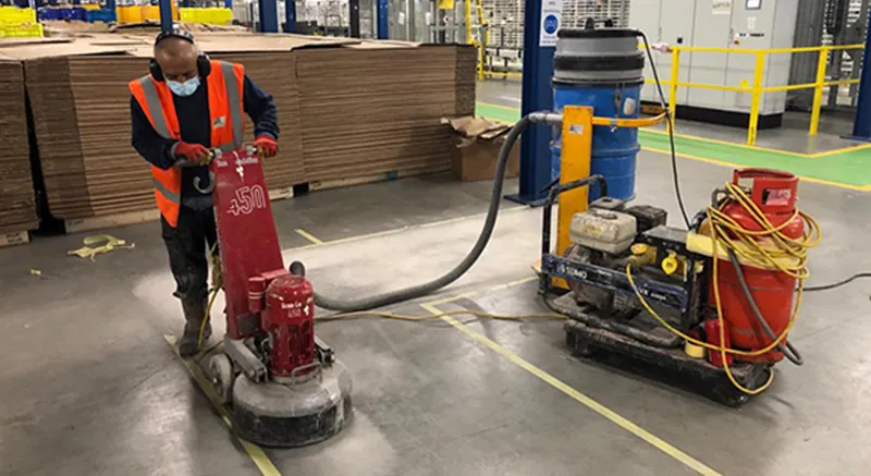 A worker wearing an orange safety vest and face mask uses a red industrial floor grinding machine in a warehouse with stacked cardboard and machinery in the background.