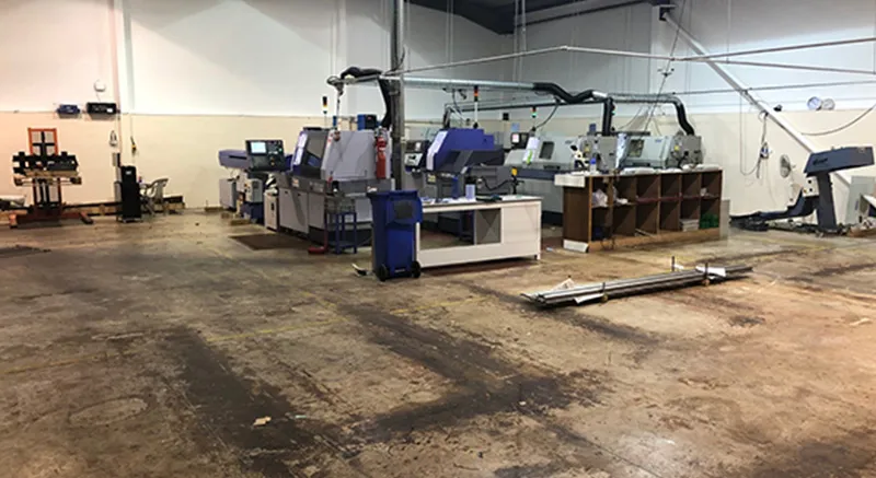 A spacious industrial workshop with large CNC machines and workstations, surrounded by empty floor space and shelves along the wall. The area appears clean and organized, with overhead lighting and visible pipes.