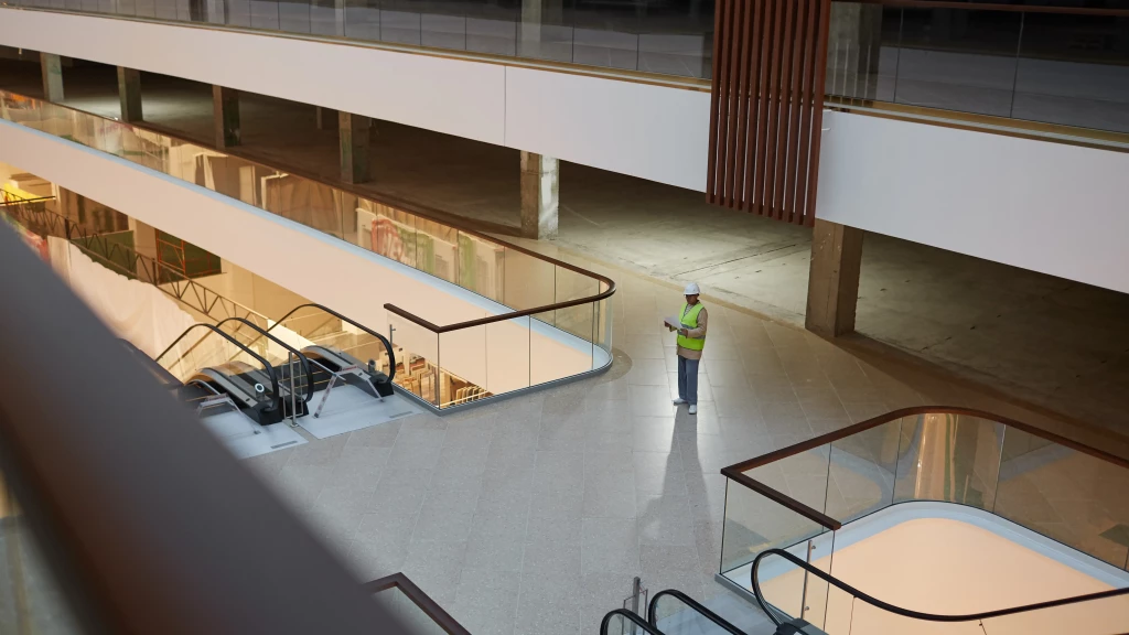 A construction worker in a yellow vest and hard hat stands alone near escalators in a large, modern, and mostly empty building with multiple floors and glass railings.