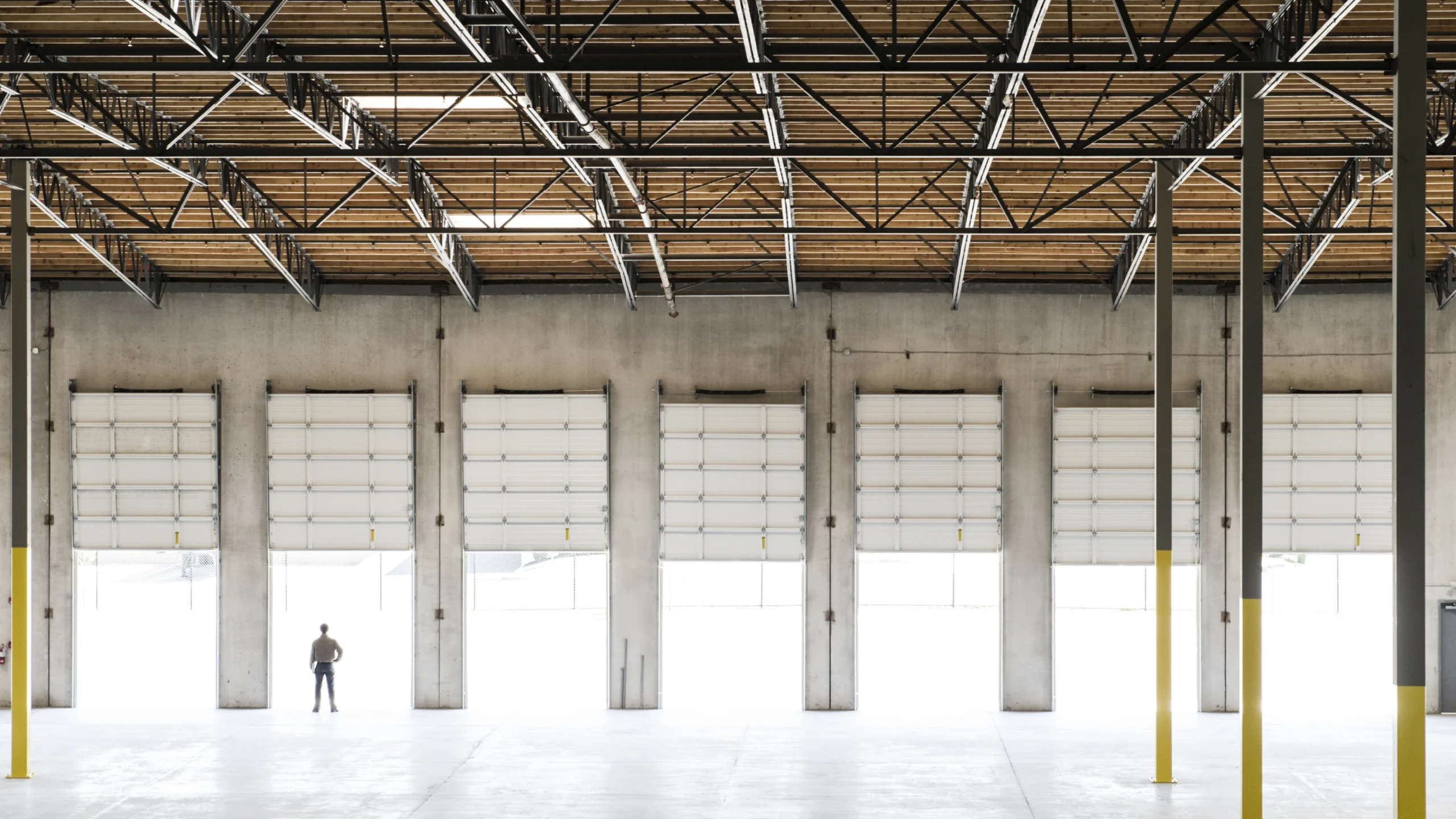 A large, empty warehouse interior with high ceilings, exposed beams, and six tall roll-up doors. A single person stands near one door, emphasizing the spaciousness and scale of the building.